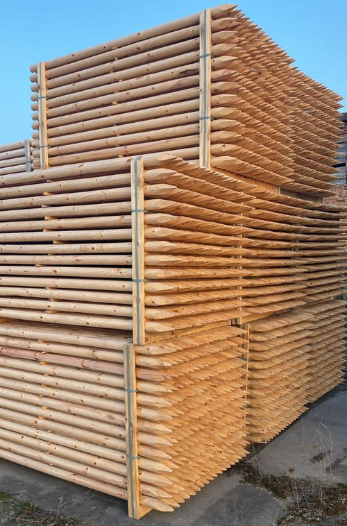 Bundled debarked pine tree stakes with milled points, stacked in the yard against a clear sky